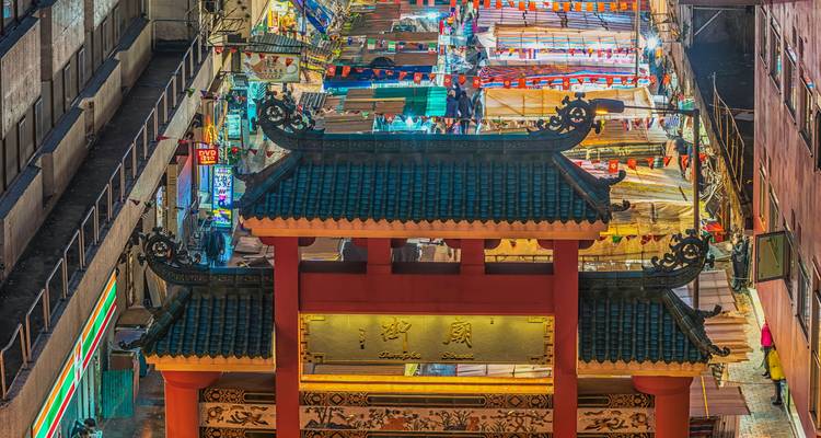 Vista nocturna hacia abajo de un mercado callejero cubierto brillantemente iluminado más allá de una puerta tradicional china ornamentada con techo de tejas verdes.