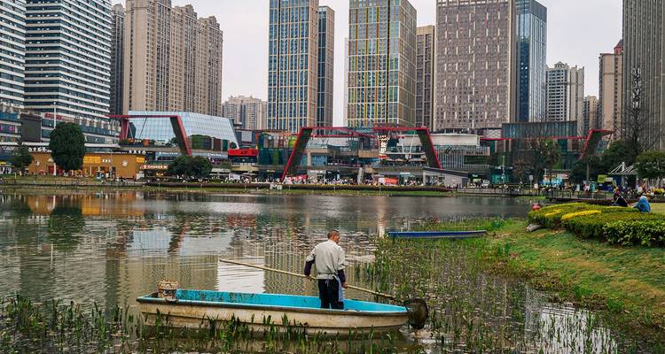 Pescador de pie en una pequeña barca en un lago urbano con rascacielos de fondo.