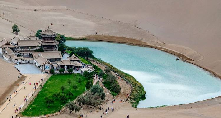 A turquoise crescent-shaped lake and temple complex surrounded by towering desert dunes