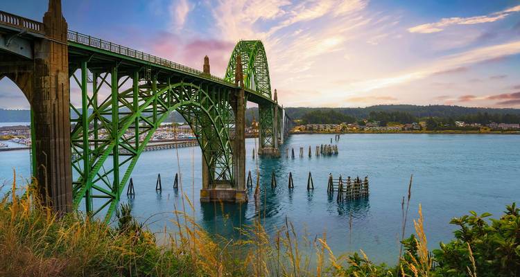 Puente de acero verde arqueado cruzando una bahía costera al amanecer colorido con aguas tranquilas abajo.