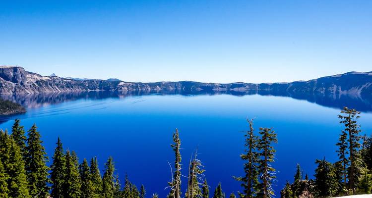 Lago de caldera volcánica de un azul brillante rodeado de acantilados escarpados y bosques de coníferas bajo un cielo sin nubes.