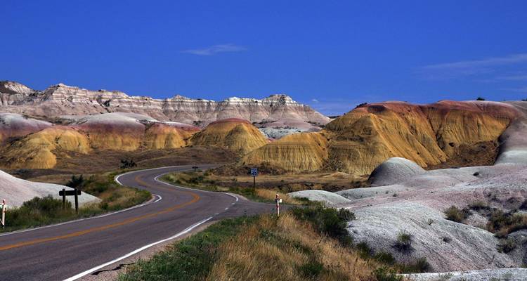 Une route sinueuse serpente à travers les buttes multicolores et les formations érodées du parc national des Badlands sous un horizon dégagé.