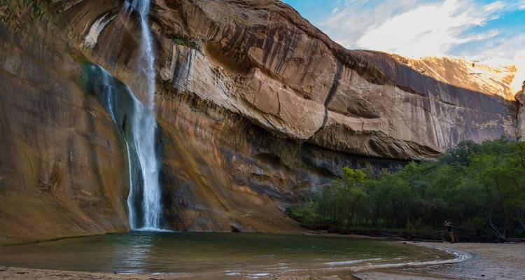 Una alta cascada cae en picada por acantilados de arenisca rayada hacia una piscina verde rodeada de árboles en la naturaleza virgen de Escalante.