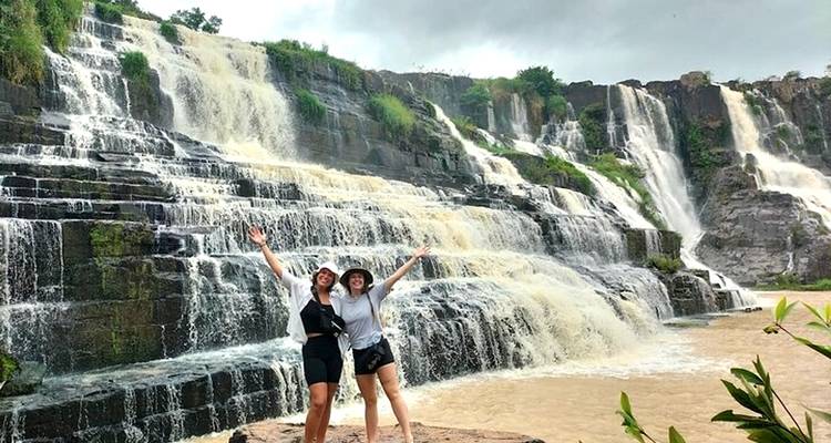Two friends raise their arms excitedly before multi-tiered waterfalls cascading over dark rock cliffs.
