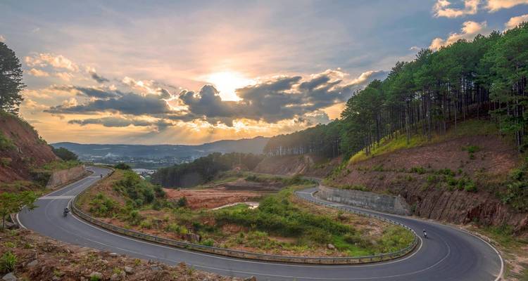 A winding mountain highway curves through pine-covered hills beneath a dramatic sunset sky.