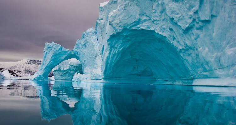 Immense arche d'iceberg turquoise se reflétant parfaitement dans les eaux arctiques calmes sous un ciel de tempête.