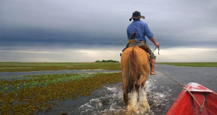 Persoon die te paard door ondiep water rijdt, gevolgd door een boot.