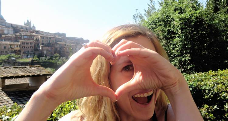 A woman making a heart shape with her hands, buildings in the background.