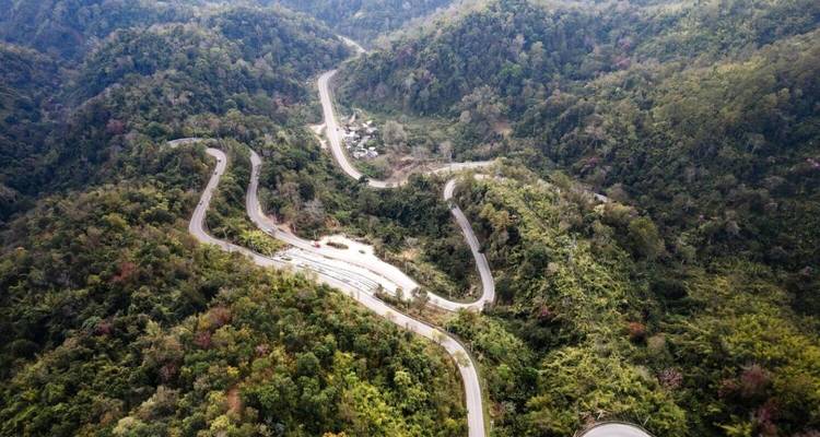 A winding mountain road snakes through thick green forested hills viewed from above.