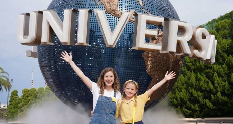 Mujer sonriente y niña pequeña con los brazos levantados frente al gigantesco globo de Universal Studios que rota y árboles verdes.