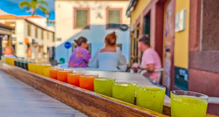 Row of colourful fruit shots in plastic cups on a wooden tray with blurred friends chatting behind