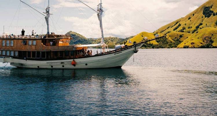 Traditional wooden phinisi sailing vessel anchored on calm turquoise waters against rolling green isles