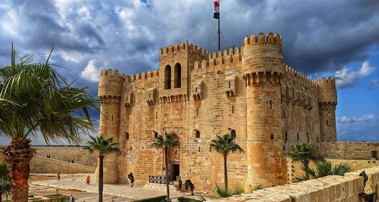 Citadel of Qaitbay with visitors and palm trees under a cloudy sky.