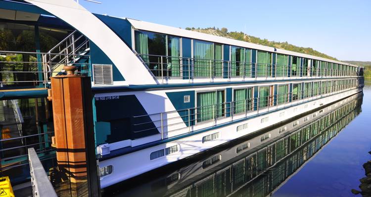 Long, modern river-cruise ship docked on a calm river beside forested hills under clear blue sky.