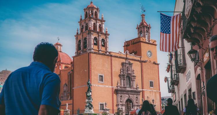 Vista a nivel de calle de una iglesia colonial de tonos cálidos con torre del reloj mientras los peatones pasan caminando bajo un cielo azul.