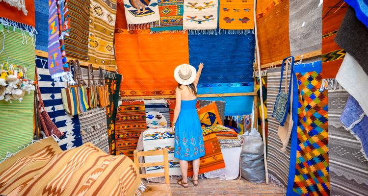 Mujer con sombrero admira alfombras tejidas vibrantes y textiles exhibidos dentro de un puesto de mercado mexicano tradicional.
