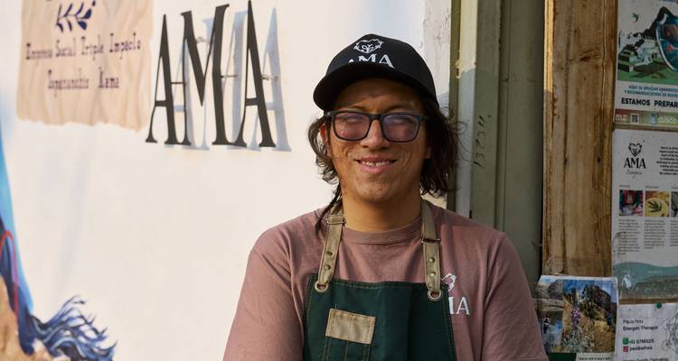 Smiling young man in apron and cap stands outside social enterprise café with AMIA sign.