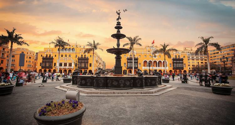 Sunset hues light Lima’s Plaza Mayor fountain surrounded by colonial yellow buildings and palm trees.