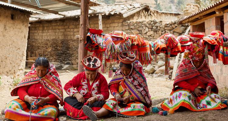 Peruvian women in vivid textiles weave seated beneath colourful yarn-covered looms in rural courtyard.