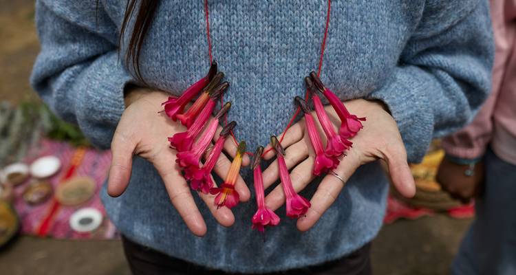 Hands display handmade necklace strung with bright pink tubular flowers against blue sweater background.