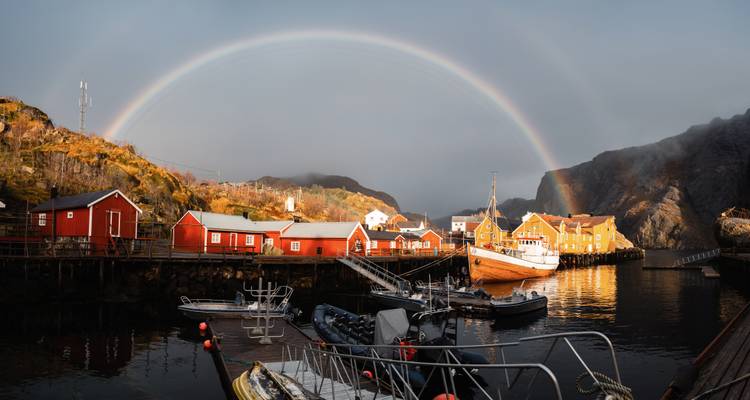 Un arco iris se forma sobre cabañas de pesca rojo reflejadas en aguas tranquilas del puerto bajo cielos brumosos.