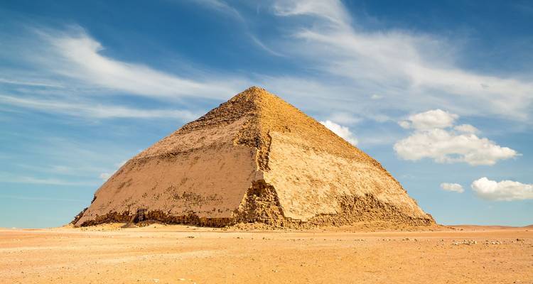The distinctive Bent Pyramid rising from the desert sands beneath a blue sky with clouds.