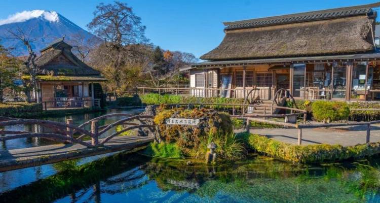 Traditionelles reetgedecktes Gebäude neben klarem Wasser mit dem Berg Fuji im Hintergrund.