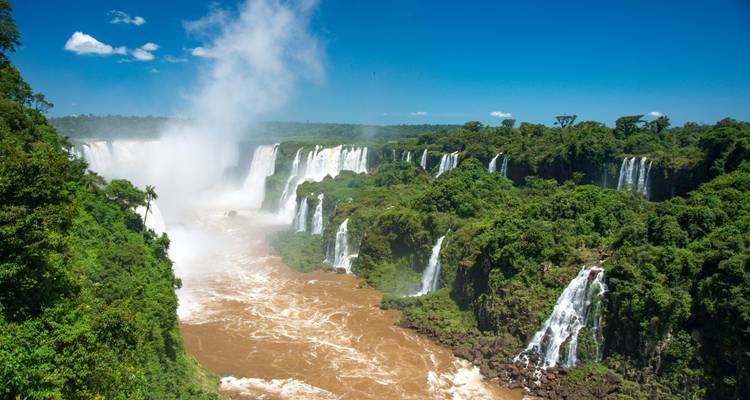 Weitblick auf die tosenden, schlammigen Iguazu-Fälle, umgeben von dichtem Dschungel unter einem klaren blauen Himmel.