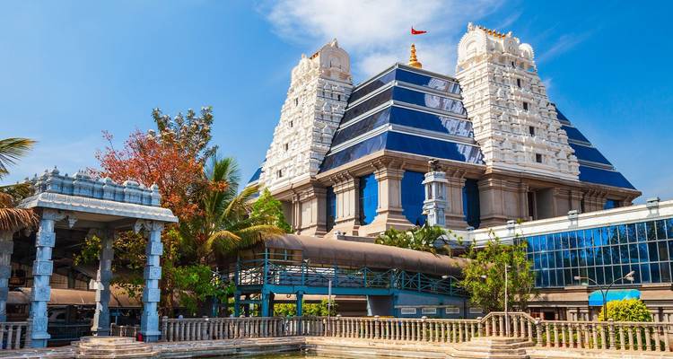 White-topped ISKCON temple with blue glass panels and a small pond in front on a bright day.