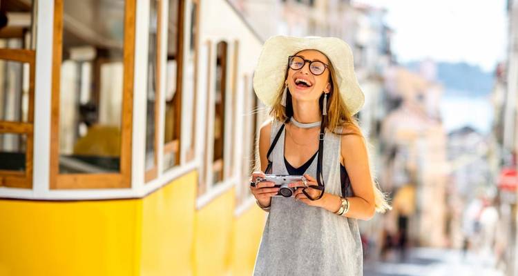 Vrouw met camera lachend naast een gele tram op een stadsstraat.