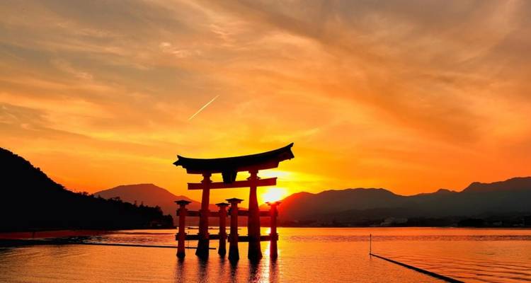 Iconische rode torii-poort lijkt te zweven op rustig water bij zonsondergang met bergen silhouet op de achtergrond.