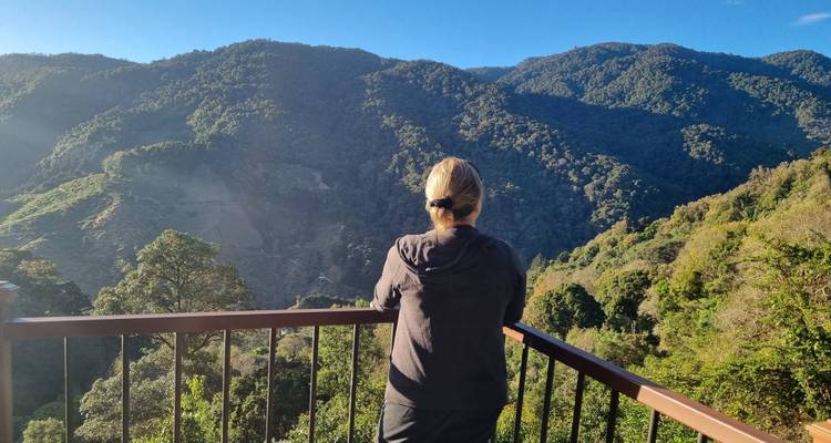 Una mujer contempla desde un balcón un valle de montaña cubierto de bosque bajo un cielo azul despejado por la mañana.