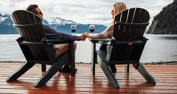 Paar sitzt auf Adirondack-Stühlen an einem See und hält Händchen.