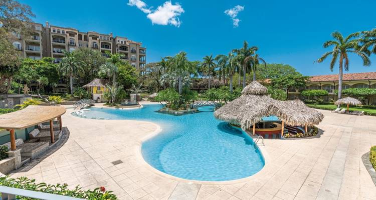 Piscina de resort tropical con lagunas de forma libre, palapas de paja, palmeras y un edificio de hotel de varios pisos más allá.