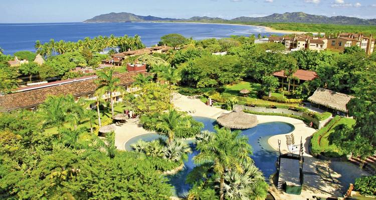 Vista aérea de un complejo turístico frente a la playa rodeado de exuberante vegetación, piscinas sinuosas y una costa que se extiende hacia el Océano Pacífico.
