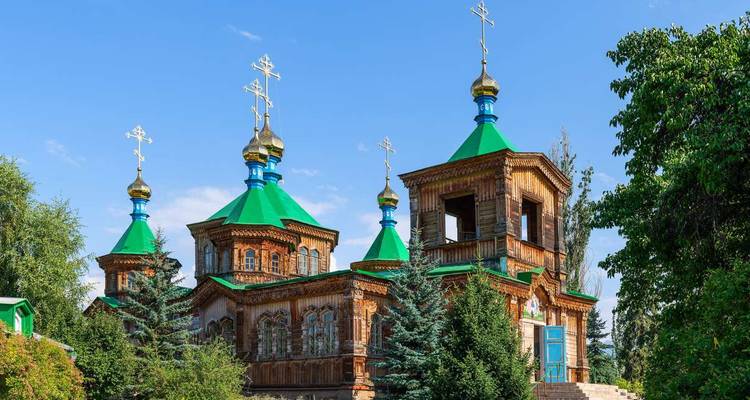 Sierlijke houten kerk met groene, gouden en blauwe koepels omringd door bomen.