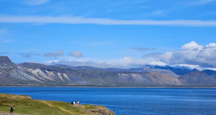 Calm fjord stretches beneath blue skies and distant mountains as a few walkers enjoy the grassy shore.