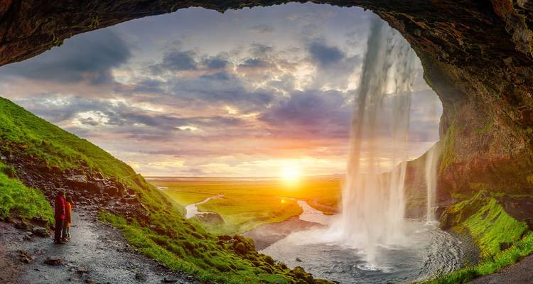 View from behind a cascading waterfall frames a glowing sunset over lush Icelandic plains.