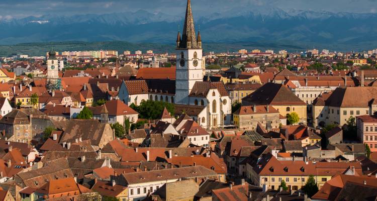 Une perspective aérienne détaillée de Sibiu montrant la flèche de la cathédrale au-dessus d'une mer de toits en terre cuite et de pics alpins lointains.