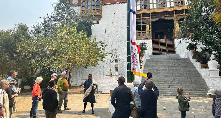 El guía habla con los turistas fuera de un templo encalado, con un colorido mástil de banderas de oración en primer plano.