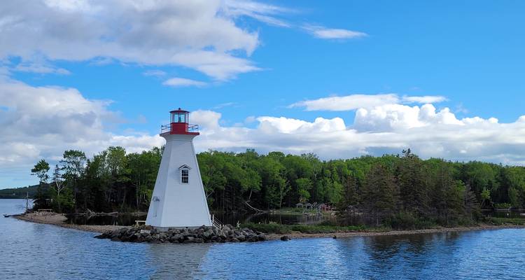 Weißer hölzerner Leuchtturm mit roter Laterne erhebt sich von einer winzigen, baumbewachsenen Insel unter blauem Himmel