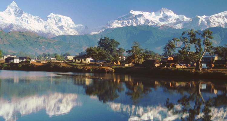 Sneeuwbedekte Himalayatoppen weerspiegelen zich perfect in een rustig meer met kleine plattelandshuisjes aan de oever.