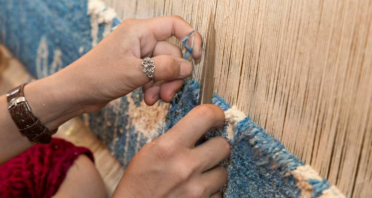 Las manos usando una cuchilla para recortar hilos azules en un telar de alfombra tradicional
