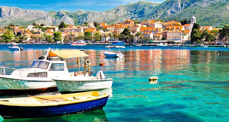 Colorful boats bob on crystal water with coastal village and mountains behind in Croatia.