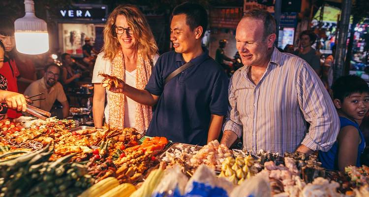 Travellers sampling street food at a lively night market stall filled with fresh produce.
