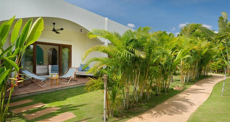 White bungalow surrounded by lush palms and a sandy path under clear blue sky.