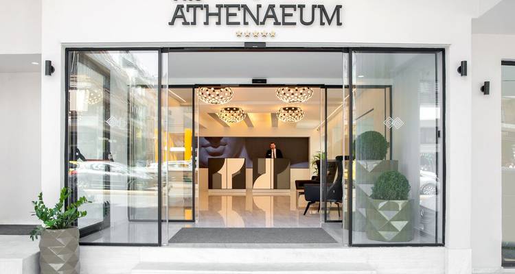 Moderne glazen ingang van The Athenaeum hotel met conciërge die achter strakke receptiebalie staat.