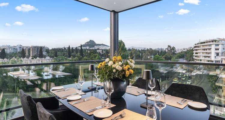Elegante eettafel op een glazen balkon met uitzicht op Athene met de Lycabettusheuvel omlijst door blauwe lucht.