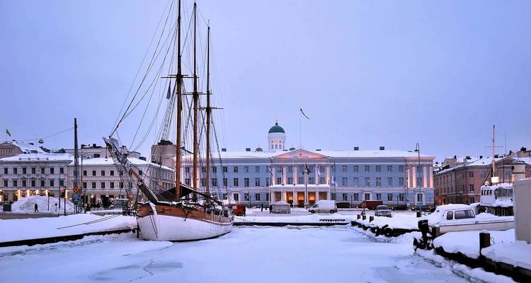Puerto cubierto de nieve en Helsinki con un barco de vela clásico y el ayuntamiento neoclásico al atardecer.
