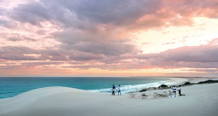 Kinematografische Ansicht über Sanddünen mit Menschen, die ein Sonnenuntergangs-Mahlzeit-Setup genießen.
